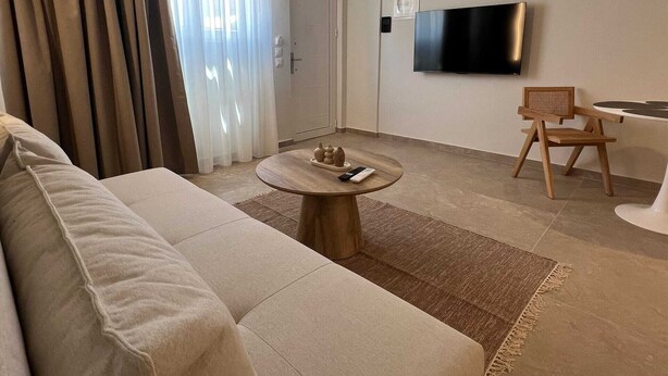 A modern living room with a sofa, a round coffee table, a TV mounted on the wall, and a dining area with a chair and table. - Ixia Cozyline Apartment - 104 (Rhodes)