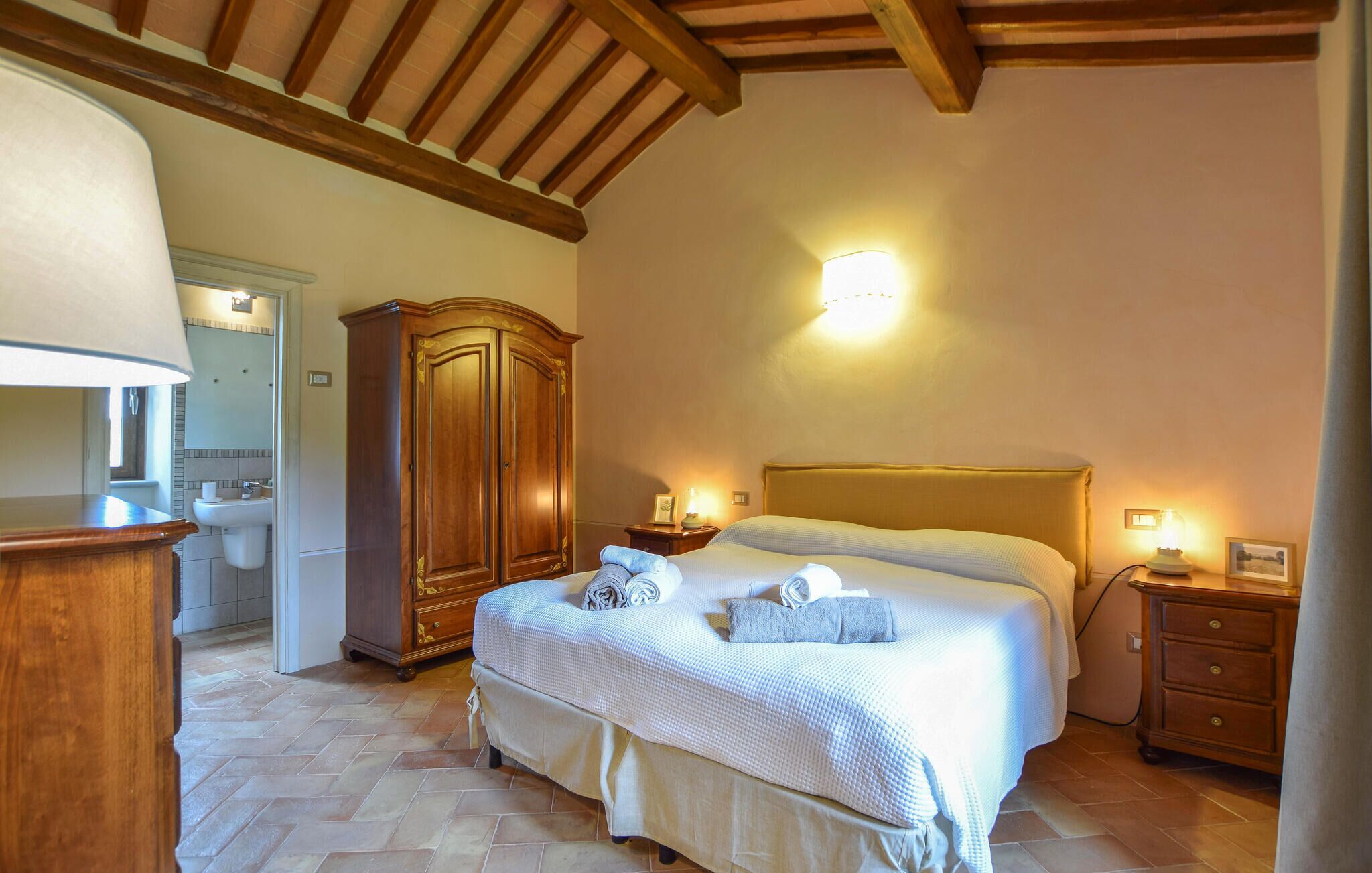 A bedroom with a bed, bedside tables, a wardrobe, a bathroom with a sink and mirror, and a wooden ceiling.