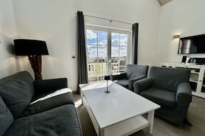 A modern living room with a white coffee table, grey sofas, a flat-screen TV, and a balcony with curtains.