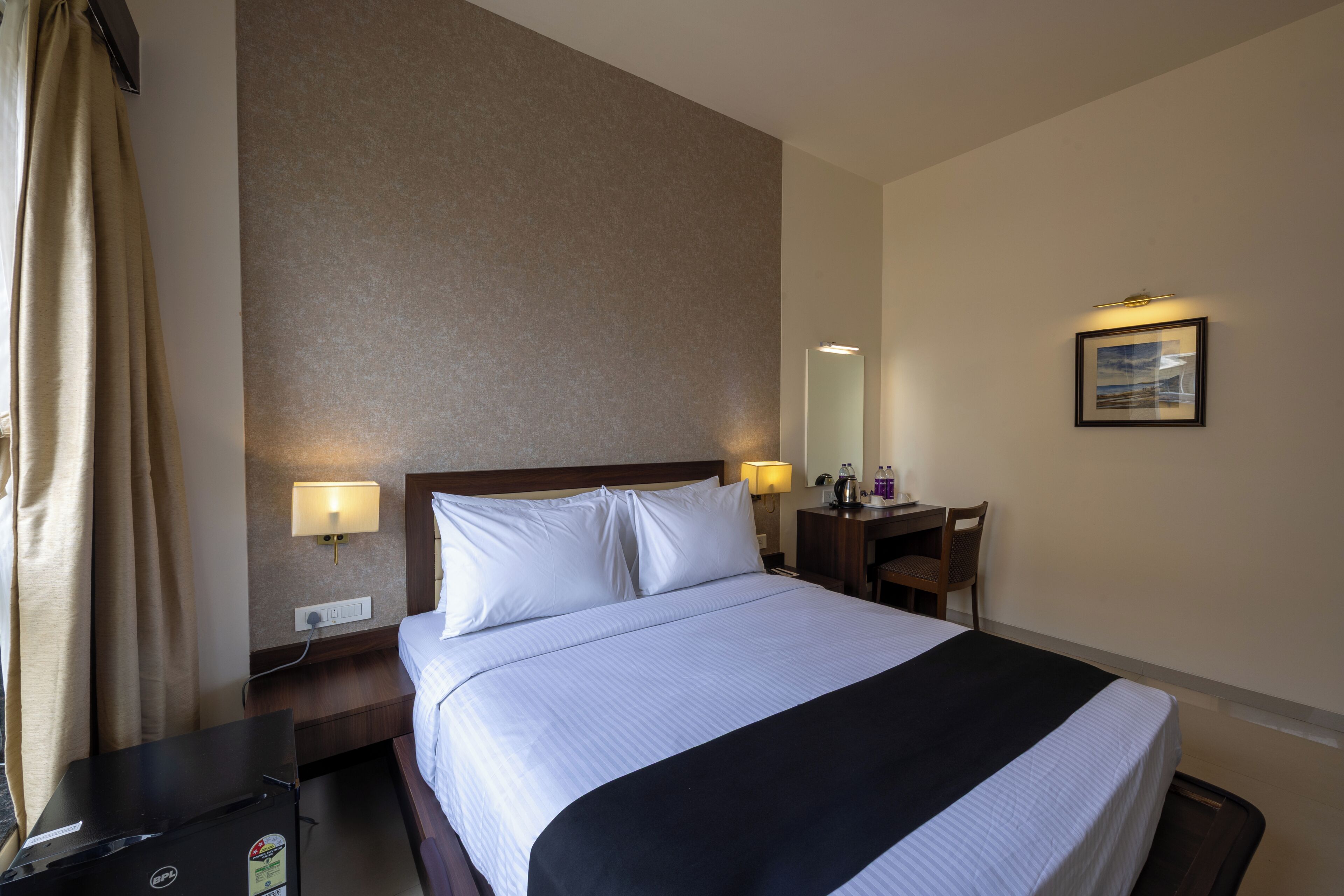 A hotel room with a bed, bedside lamps, a desk, a chair, a mirror, and a wall-mounted picture.