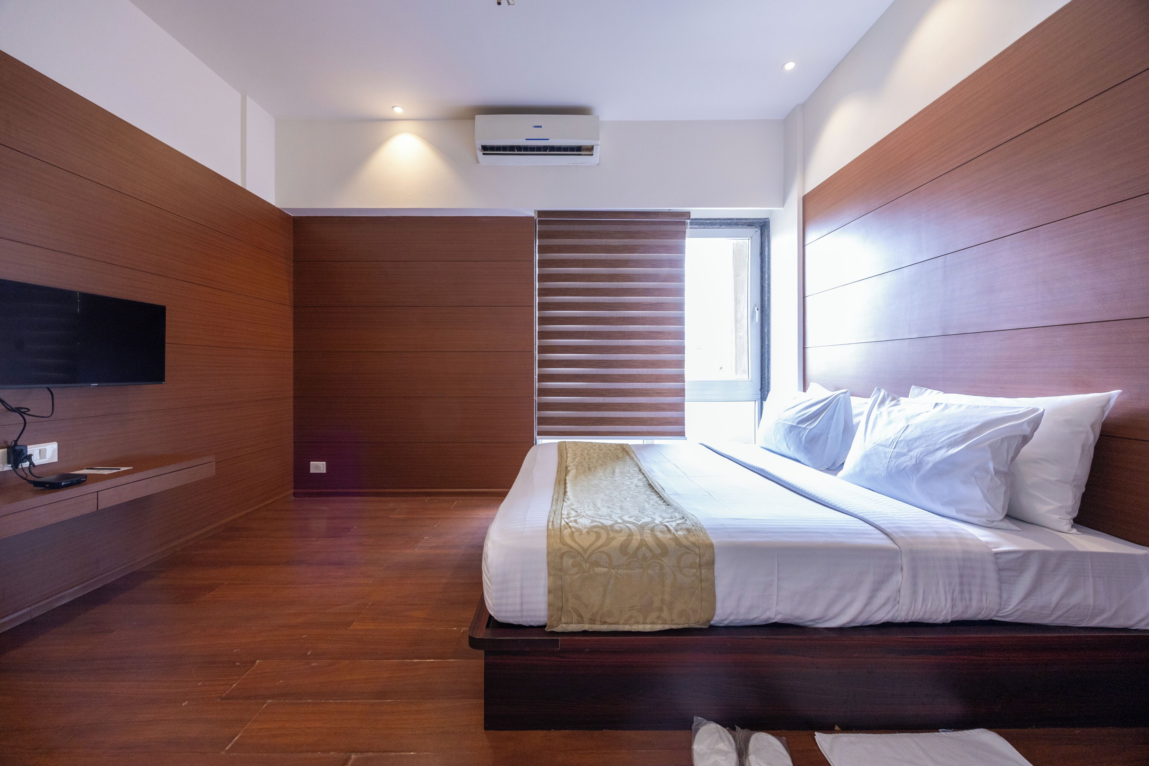 A modern hotel room with a wooden floor, a large bed with white linens, a mounted TV, and a window with blinds.