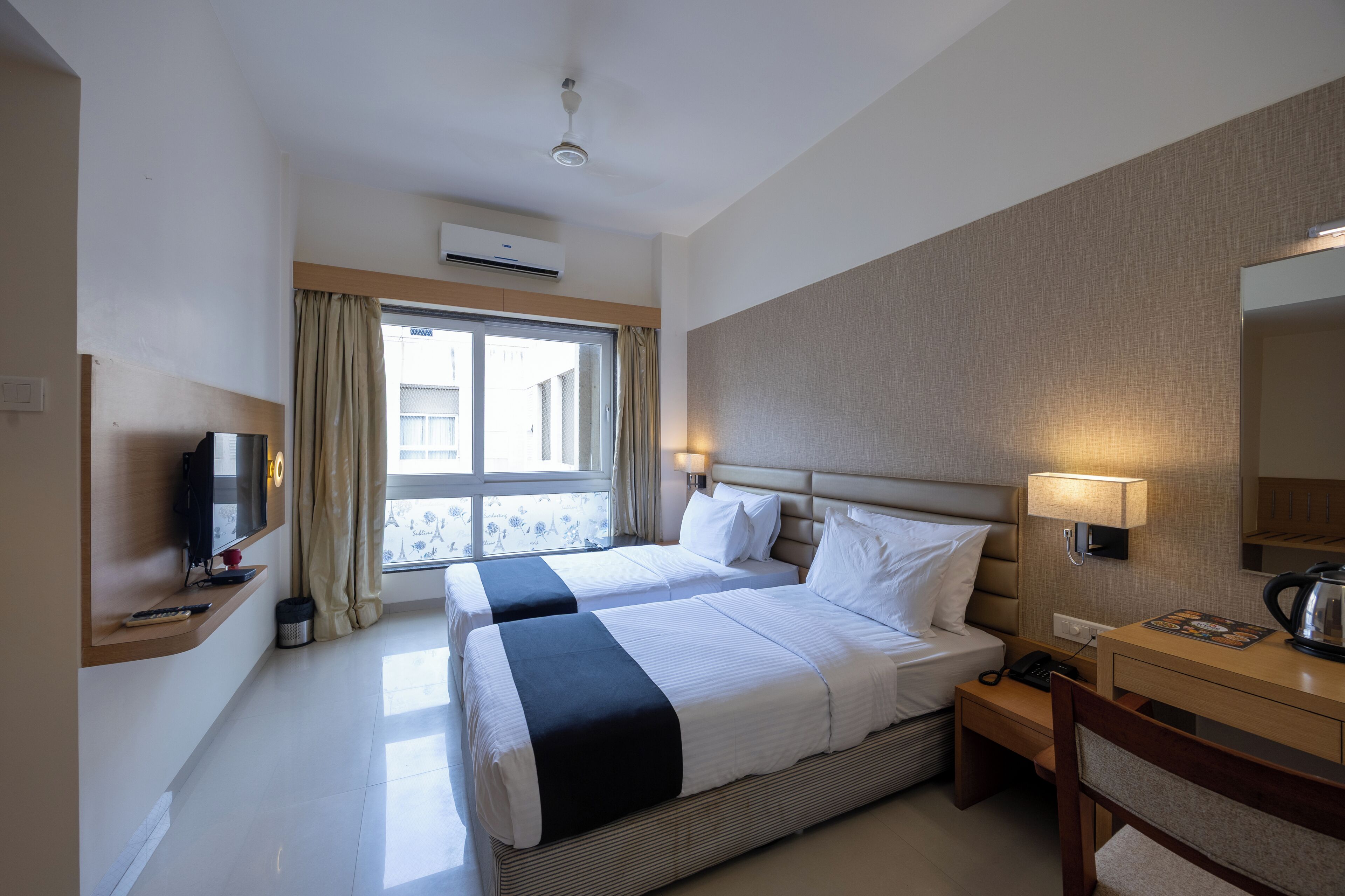 A hotel room with two beds, a TV, a desk with a lamp, and a window with curtains.