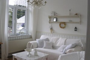 Ein helles Zimmer mit einer Couch, einem Tisch mit weißer Tischdecke, einem Fenster mit Aussicht und einer Kristallleuchter.