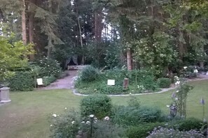 Ein Garten mit verschiedenen Pflanzen und Bäumen, einem Weg und einer kleinen Struktur im Hintergrund.