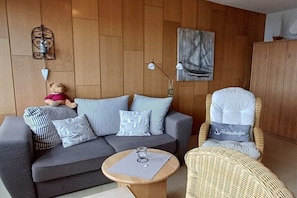 A cozy room with a grey sofa, a wicker chair, a small round table, and a wall-mounted lamp. - Trumann "Laughing Gull" (Fehmarn (Südstrand))