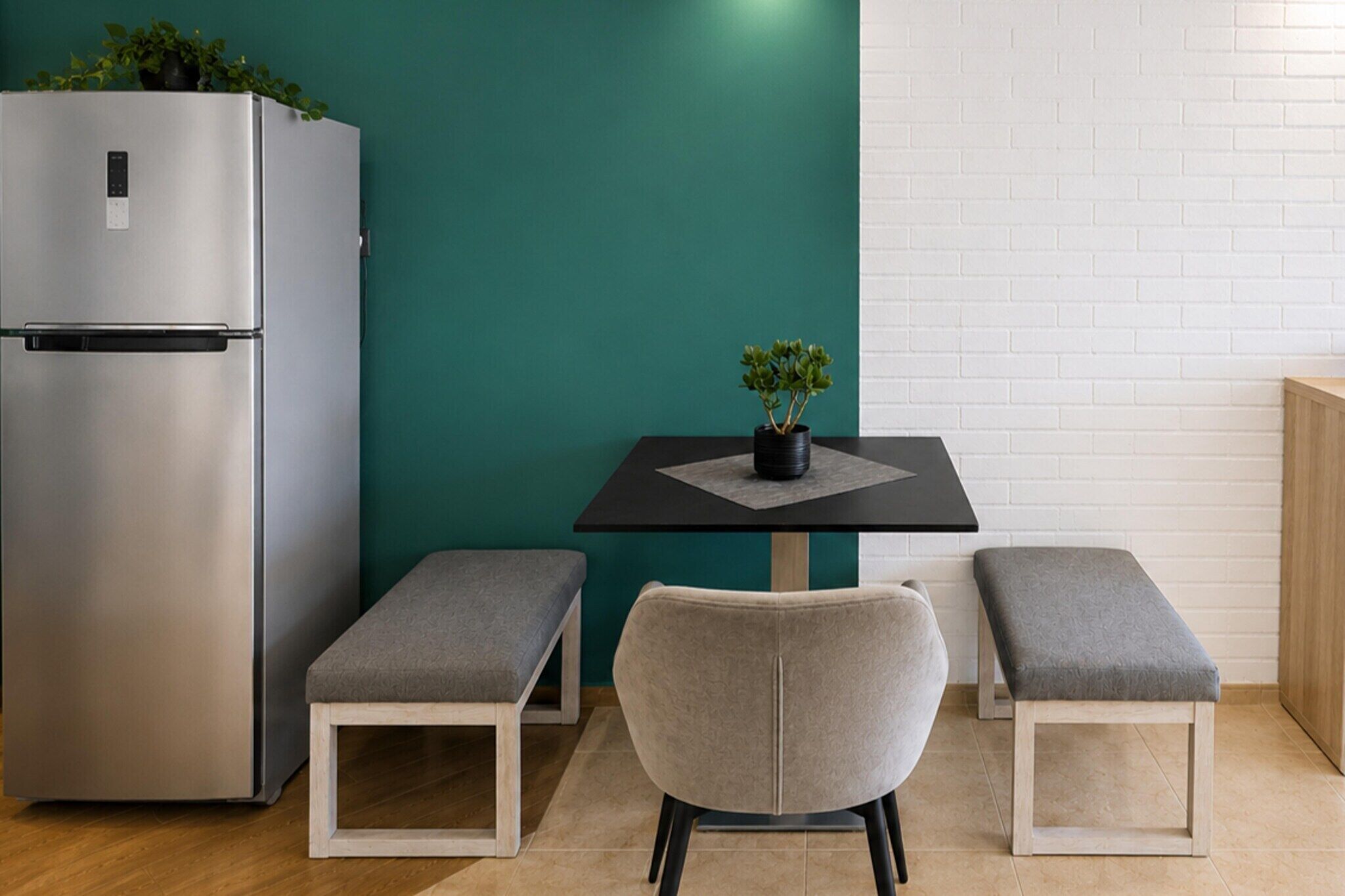 A compact kitchen with a stainless steel refrigerator, a small table with a potted plant, and two grey cushioned benches.