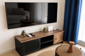 Ein modernes Wohnzimmer mit einem großen Flachbildfernseher an der Wand, einem Holzkonsolentisch mit Fernbedienung und einer kleinen Pflanze sowie einem runden gewebten Hocker.