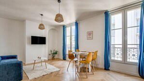 Una habitación amplia con zona de comedor, televisor montado en la pared y ventanas grandes con cortinas azules.