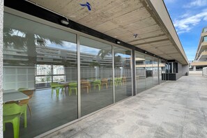Ein modernes Gebäude mit großen Glasfenstern, einer Außenbestuhlung mit grün-gelben Stühlen und einem Betonboden.