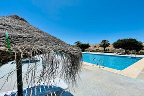 A swimming pool with a thatched umbrella, lounge chairs, and a clear blue sky. - Max Apartment with communal garden and pool (Roquetas de Mar)