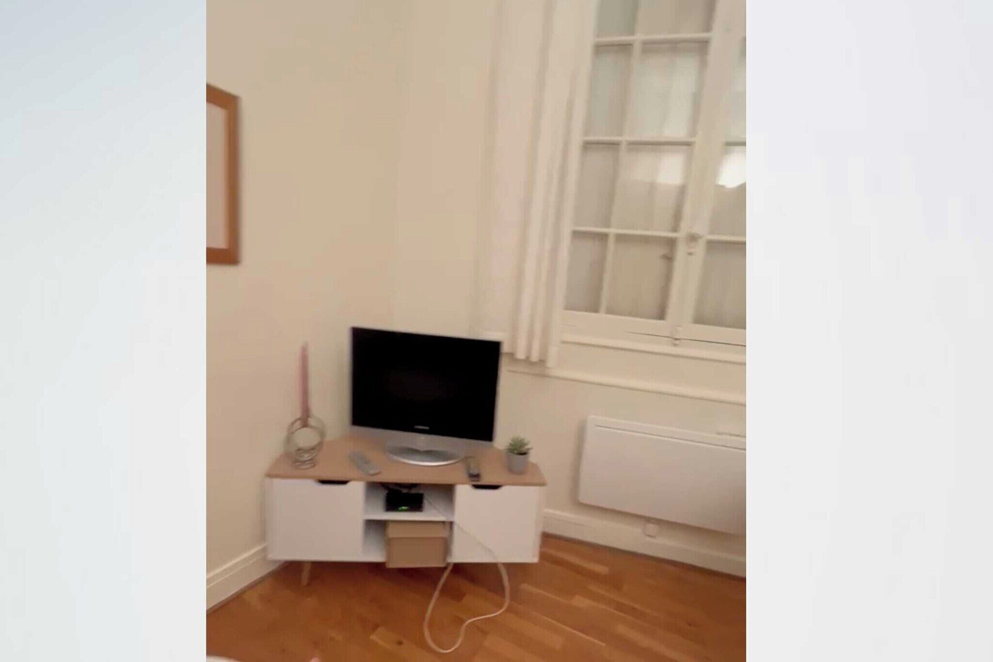 A small, neatly organized room with a TV on a wooden stand, a white radiator, and a window with white curtains.