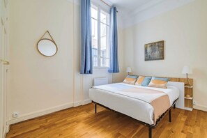 Un dormitorio con cama, mesita de noche, lámpara, ventana con cortinas, espejo en la pared y un cuadro.