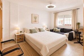 Ein Schlafzimmer mit einem großen Bett, Nachttischen, einem Sessel, einer Couch, einem Fenster mit Vorhängen und einem Deckenventilator.