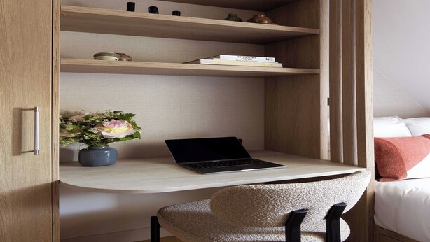 Una habitación con escritorio de madera integrado, una computadora portátil, un florero con flores y una cama con ropa de cama blanca y rosa.