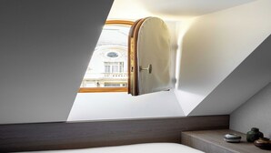 Una habitación con techo inclinado, una cama, una mesita de noche con un jarrón y vistas a un edificio a través de una ventana.