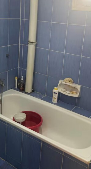 A bathroom with blue tiles, a white bathtub, a red bucket, and toiletries on the wall. - Bed and breakfast bejaia (Béjaïa)