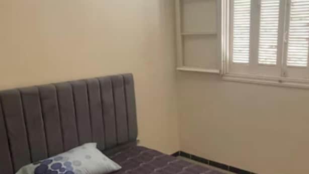 A bedroom with a bed, a pillow, a blanket, a window with blinds, and a wall-mounted shelf.