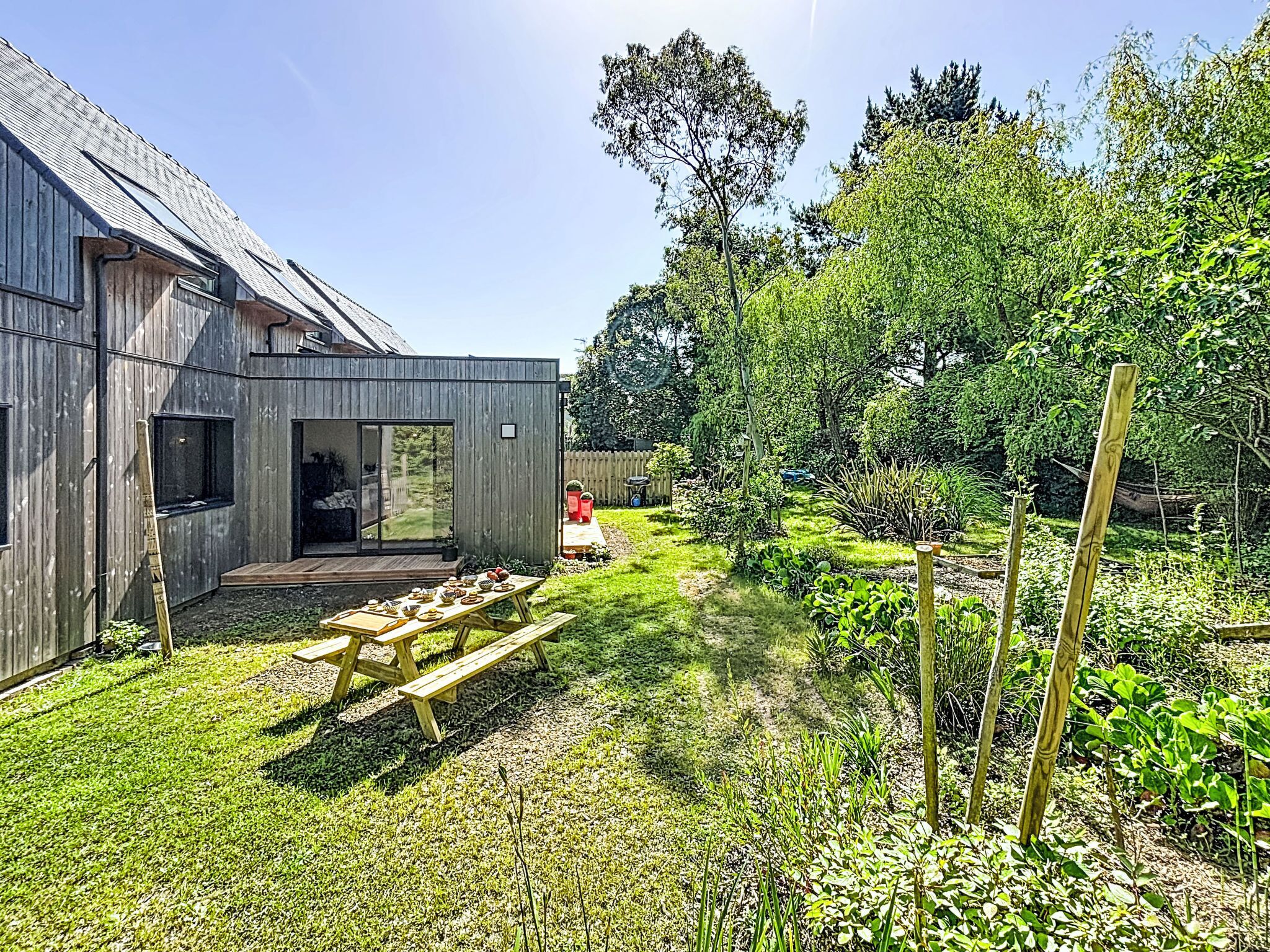 A wooden building with a patio, a table with chairs, and a garden with various plants.