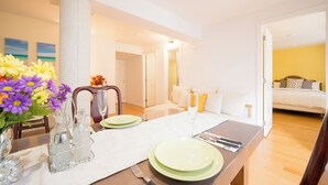 Spisestue med et bord dækket til to personer, en vase med blomster og udsigt til et soveværelse med seng og et natbord.