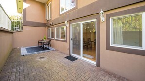 En overdækket terrasse med et bord og stole, en skydedør der fører til et spiseområde indendørs, og et lille udendørs loungeområde med et bord og stole.