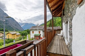 Un balcón de madera con vistas a un paisaje montañoso, un edificio blanco con techo verde y una silla roja.