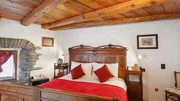 A wooden-framed bed with a red blanket, a stone wall, and a wooden nightstand with a lamp.