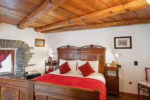 Una cama con estructura de madera, cubierta con un edredón rojo, una pared de piedra y una mesita de noche de madera con una lámpara.