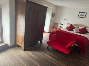 Una stanza con un armadio in legno, un divano rosso, un letto con cuscini rossi e bianchi, e un pavimento in legno.