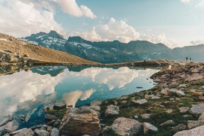 Eine Berglandschaft mit einem klaren See, der Himmel und Berge spiegelt, felsigem Gelände und einigen Personen in der Ferne.