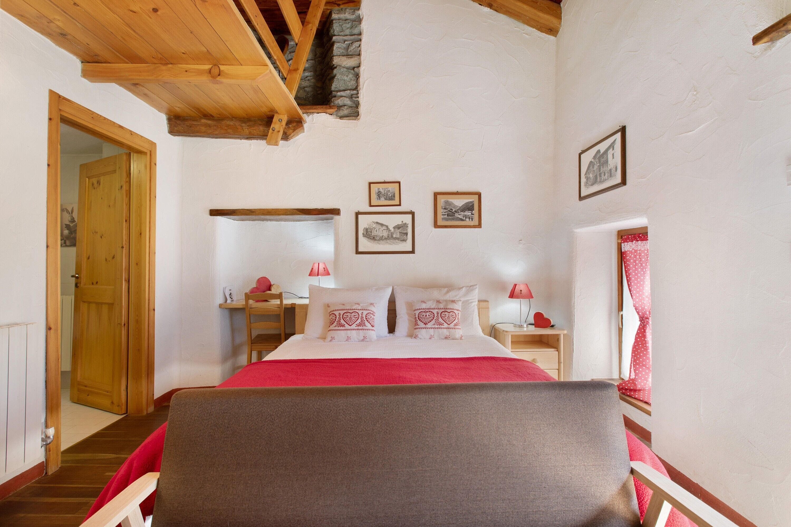 Une chambre avec un plafond en bois, un lit avec une literie blanche et des touches de rouge, une table de chevet avec une lampe, et une fenêtre avec des rideaux rouges.