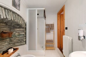 Une salle de bain avec un mur en pierre, une étagère en verre, une étagère en bois avec des serviettes, un radiateur blanc et une porte en bois.
