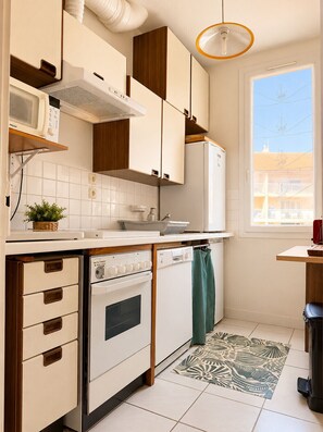 Eine kompakte Küche mit weißen Schränken, einer Mikrowelle, einem Backofen und einem Geschirrspüler. Ein Fenster bietet Blick auf umliegende Gebäude, und ein gemustertes Teppich liegt auf dem Boden.