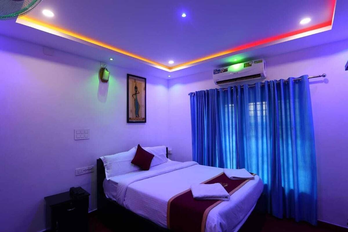 A hotel room with a bed, a nightstand, an air conditioning unit, and a window with blue curtains.