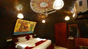 A bedroom with a bed, a wooden wardrobe, a mirror, and a wall with a colorful painting. - Wayanad Cave N Pillar Resort (Krishnagiri)