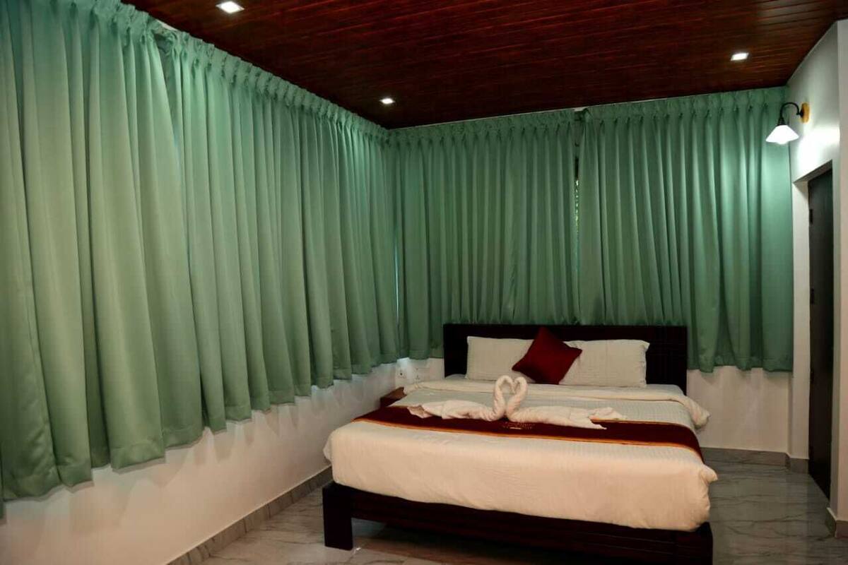 A neatly made bed with a heart-shaped towel, a dark headboard, and green curtains.