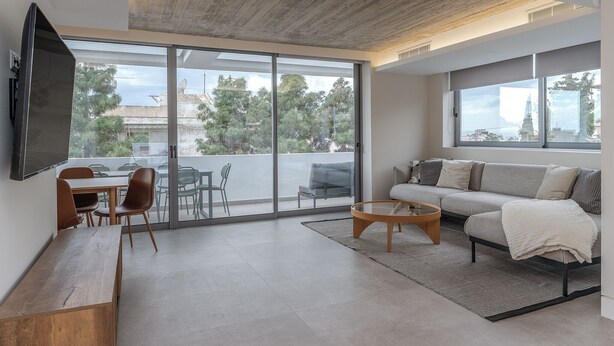 A modern living room with a sofa, coffee table, and TV, large sliding glass doors to a balcony, and a wooden ceiling. - Sea View Riviera Maisonette with Large Terraces (Voula)