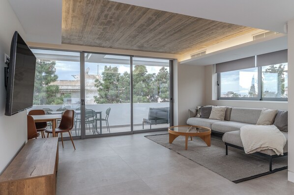 Ein modernes Wohnzimmer mit einer Couch, einem Couchtisch und einem Fernseher, großen Schiebeglastüren zu einem Balkon und einer Holzbalkendecke.