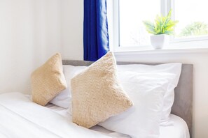 A bed with two beige pillows and a white pillow, a window with a plant, and blue curtains.