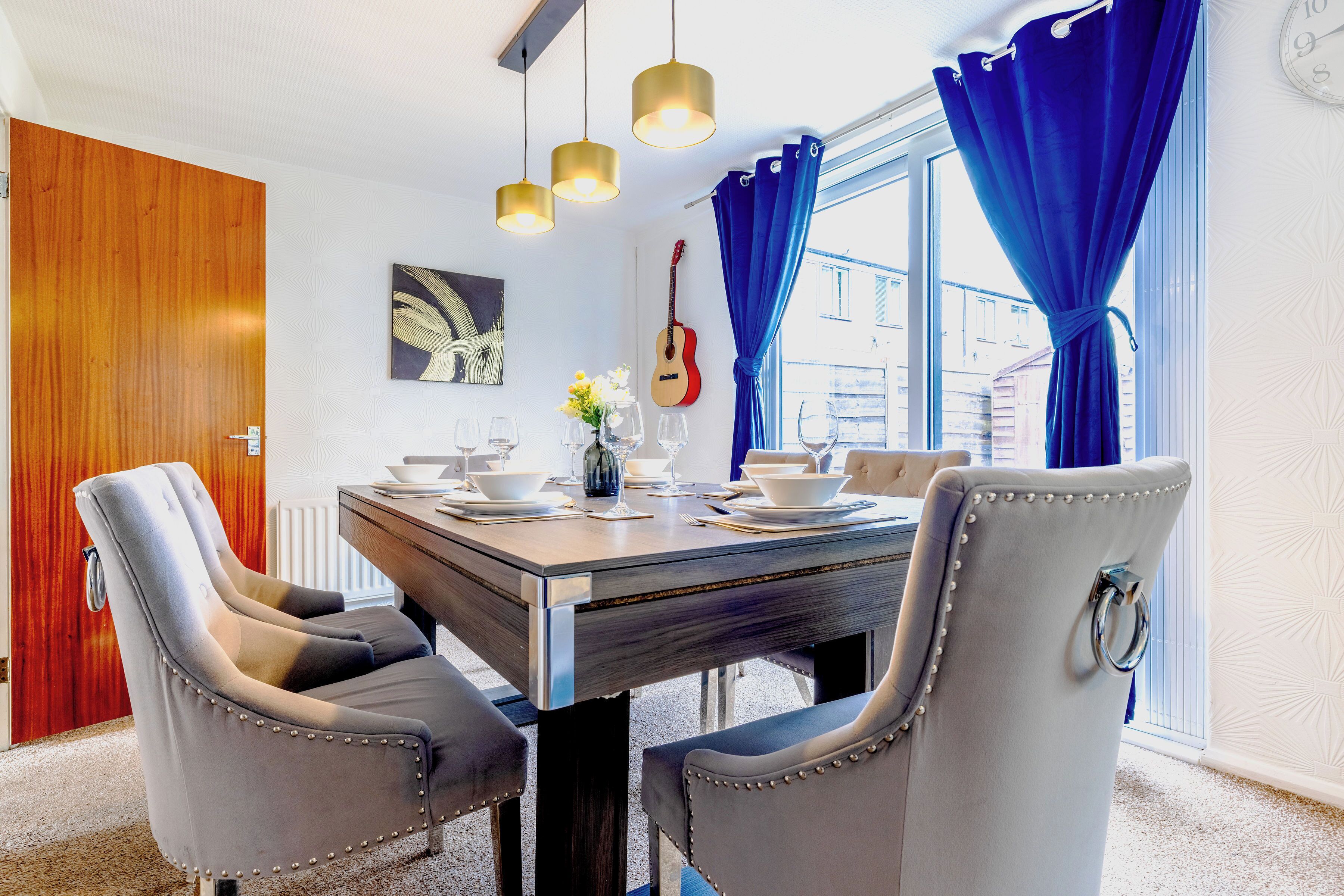 Un coin repas avec une table en bois, des chaises à revêtement gris, une guitare et une fenêtre ornée de rideaux bleus.