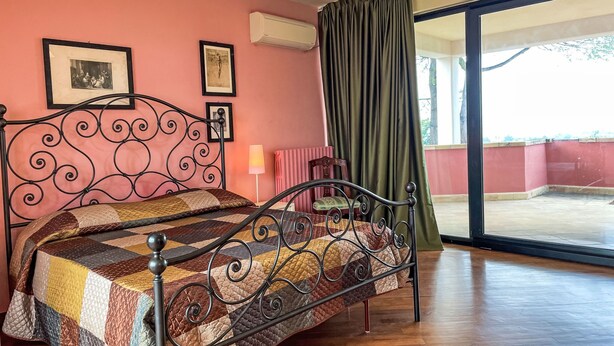 A bedroom with a metal bed frame, a quilted bedspread, a window with a view, and a wall-mounted air conditioner. - Vacation home Vidal by Interhome (Spoltore)