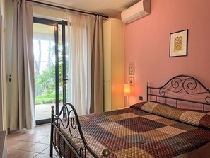 Een kamer met een bed, een deur die leidt naar een balkon, een airconditioningapparaat en een aan de muur hangend schilderij.