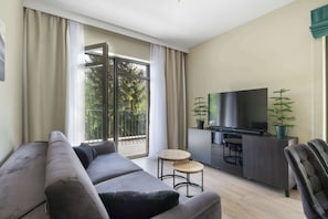 Un salon moderne avec un canapé, une petite table ronde, un téléviseur et un balcon donnant sur la verdure.