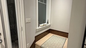 En hall med en dörr, ett fönster med en hylla, en mönstrad matta och en trappa.