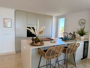 A modern kitchen with a central island, bar stools, a potted plant, and a decorative clock.