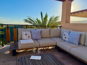 Ein beiges Ecksofa mit gemusterten Kissen, ein Holztisch mit zwei WeinglÀsern und im Hintergrund eine Aussicht auf Palmen und Berge.
