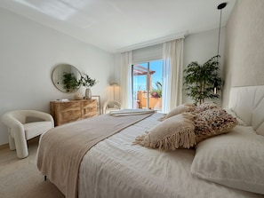 Une chambre avec un grand lit, une commode en bois, un miroir rond et une vue sur l’extérieur.
