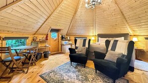 Un intérieur chaleureux avec un haut plafond en bois, un coin repas avec des chaises et une table, un canapé avec des coussins et un petit coin cuisine équipé d’appareils électroménagers.