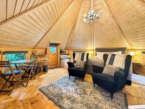 Ein gemütlicher Innenraum mit einer hohen Holzbalkendecke, einem Essbereich mit Stühlen, einer Couch und einer kleinen Küchenzeile.