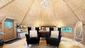 A cozy cabin interior with a high wooden ceiling, two black armchairs with white cushions, a wooden dining table, a white vanity with a mirror, and a small kitchenette with a microwave. - Willow Lodge at Aos Sí (Ballachulish)