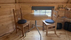 A wooden cabin interior with a round table and two chairs, a window, and a shelf with items. - Willow Lodge at Aos Sí (Ballachulish)
